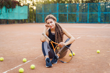 Portrait of a beautiful young woman sitting near a net tennis court among tennis balls outdoors. Sportswoman resting on a tennis court. Posing dressed in stylish sportswear