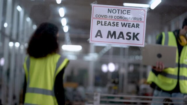 Woman Factory Worker Holding The Signage Of Wear A Face Mask To Protect Pollution And Covid 19 Or Coronavirus Pandemic. Label Attached On Clear Glass Wall. New Normal Life In Workplace Industrial.