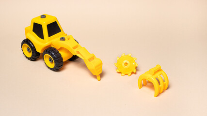 Obraz premium Children's plastic toy on a beige background - a yellow tractor with different nozzles drill, saw on wood, a wood loader, a bucket excavator. Construction equipment for children.