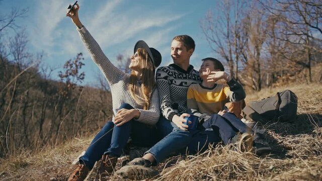 Slow-motion cinematic footage. A happy young family of mother father and little son are taking a selfie with a smartphone outdoors on a sunny spring day.