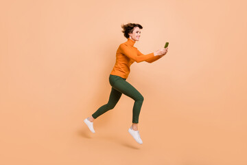 Fototapeta na wymiar Full length photo of funny cute young lady wear orange turtleneck jumping typing modern device isolated beige color background