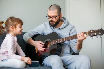 Little girl showing her father a song on a phone while he is playing guitar