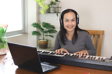 Asian woman wearing headphones playing  electric piano with  computer notebook on table  smiling...