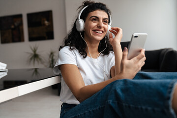 Cheerful brunette woman in headphones using mobile phone while sitting