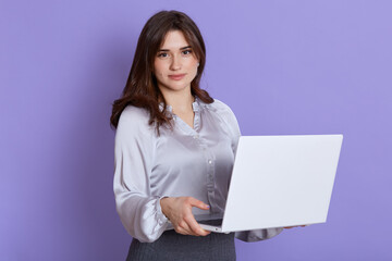 Young woman with dark hair using laptop and smiling isolated over lilac background, looking at camera, wearing elegant blouse and skirt, beautiful female with portable computer indoor.