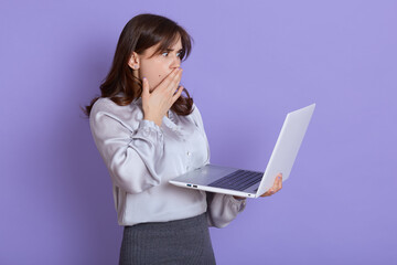 Attractive shocked businesswoman with laptop in hands posing isolated over lilac background, wearing elegant attire, has unexpected trouble with portable computer or her work, covers mouth with palm.