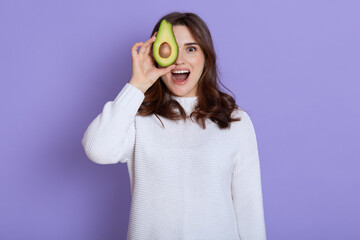 Fototapeta premium Happy excited young woman holding avocado half, covering eye with healthy fruit and keeping mouth opened, yelling something while standing against lilac wall, dresses white jumper.