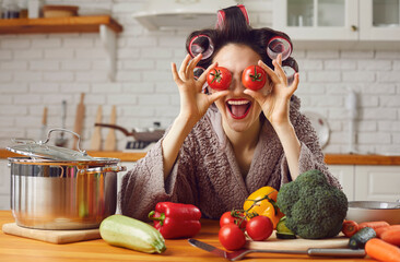 Happy positive smiling young woman having fun while cooking healthy vegetable meal at home. Funny cheerful housewife in hair curlers sitting at kitchen table, covering eyes with tomatoes and laughing