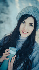 young woman in a knitted sweater and a warm hat in winter weather in a snowy pine forest with a thermo mug in her hands