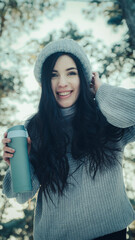 A young brunette woman with long hair in a warm knitted sweater and a winter hat with a scarf holds a thermo mug in her hands. Bottom view of the trees in the winter forest.