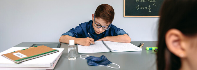 Boy writing in his notebook at school with mask and hand sanitizer on the desk