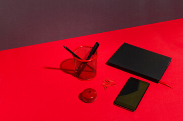 Black empty screen of phone, notebook, pen holder, pensil,pen, sharpener on the red desk against dark wall