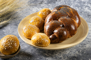 A loaf topped with poppy seeds and sesame seed burger buns lie on a bamboo platter, with wheat spikelets on the side. Bread products. Home cooking..