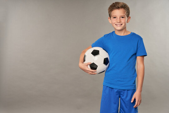 Cheerful boy with soccer ball standing against gray background