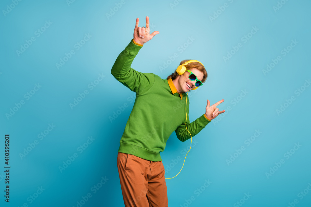 Poster photo of young happy positive smiling man in headphones and sunglasses show rock n roll sign isolate