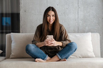 Cheerful brunette nice girl smiling and using mobile phone on sofa