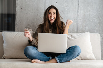 Delighted girl winking and holding credit card while using laptop at home