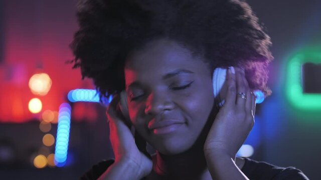 Portrait of 20s african black woman listen to music wearing headphones,color neon lights in the background,curly afro hair american girl dancing slow motion camera in a dark room colorful led lighting