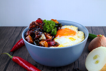 Rice in a bowl with sunny side up and fried chicken with black pepper sauce. Wooden background, chilies and onions.