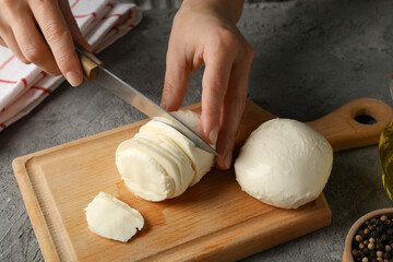 Woman slicing mozzarella on wooden board on gray table, close up