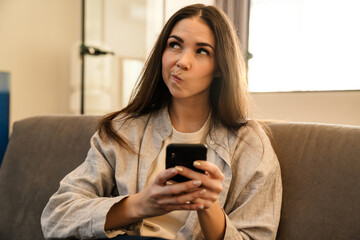 Fototapeta na wymiar Perplexed young woman using smartphone while sitting on sofa indoors