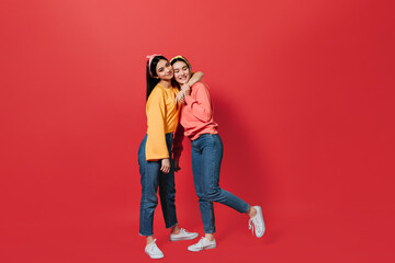 Full-length shot of girls in jeans and sweatshirts hugging on red background