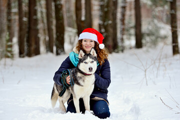 Fototapeta na wymiar Young beautiful girl with a Husky dog in the winter forest. She sits and hugs her dog. She looks into the camera lens. Horizontal orientation.