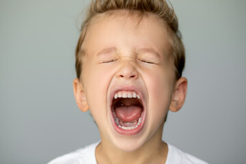 little boy screams with closed eyes. Isolated young man on gray background opened his mouth wide