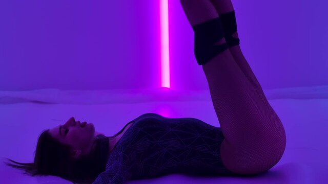 Sexy woman in high heels dancing modern plastic choreography with strip dance elements. Striptease dancer moves erotically in dark studio against backdrop of bright neon lights. Close up. Slow motion.