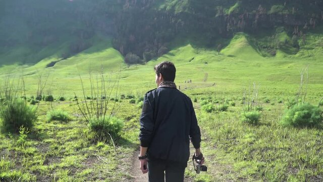 Man walking on beautiful meadow landscape at foot of Bromo Mountain National Park at Teletubbies hill (Bukit Teletubbies Bromo) and holding a camera, at East Java, Indonesia. Wide shot.