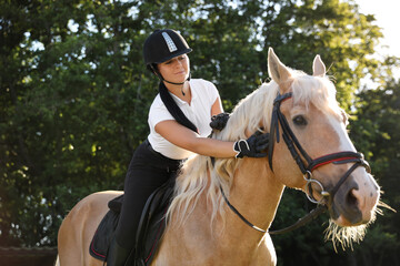 Young woman in equestrian suit riding horse outdoors on sunny day. Beautiful pet