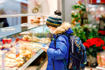 Kid boy wearing medical mask buy bread and pastry for school lunch in bakery. Child with backpack...
