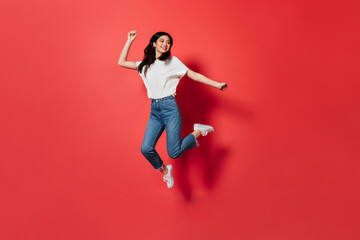 Fototapeta na wymiar Mischievous girl in white T-shirt and jeans jumping on red background