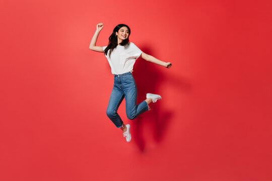 Mischievous girl in white T-shirt and jeans jumping on red background