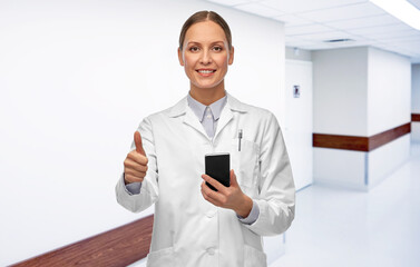medicine, profession and healthcare concept - happy smiling female doctor or scientist with smartphone showing thumbs up over hospital corridor background