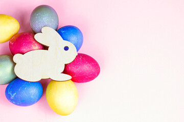 Top view flat lay funny wooden Easter Bunny with colorful eggs as symbols of a holiday with copy space on light pink background.