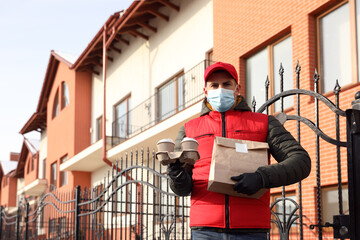 Courier in medical mask holding takeaway food and drinks near house outdoors. Delivery service during quarantine due to Covid-19 outbreak