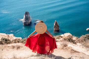 A girl with loose hair in a long red dress descends the stairs between the yellow rocks overlooking the sea. A rock can be seen in the sea. Sunny path on the sea from the rising sun