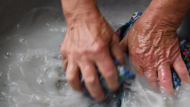 Senior hands of mixed-race 80s woman crumple and wash colorful clothes in old basin. Obsolete way to clean laundry instead of using washing machine. Wrinkled hands handwash textile in soap water