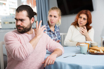 Upset young man sitting at table after quarrel and girl with mother on background at home