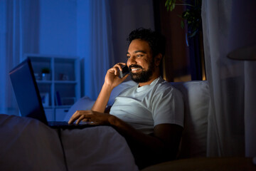 technology, remote job and people concept - happy smiling young indian man with laptop computer calling on smartphone in bed at home at night