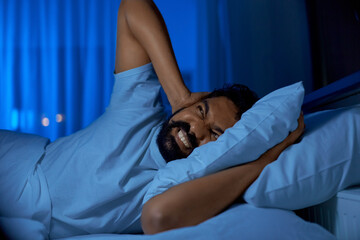 people, bedtime and rest concept - annoyed indian man lying in bed at home and covering ears at night