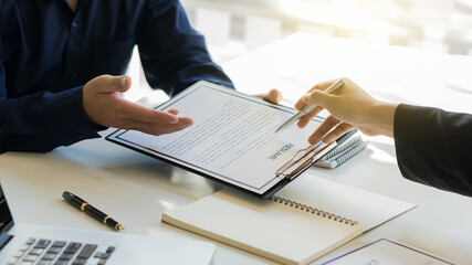 The interviewer asks the applicant to fill out his resume on paper, pointing out papers on the desk, laptop, and notebook.