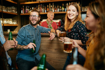 Group of cheerful friends standing near bar counter, drinking beer and chatting.