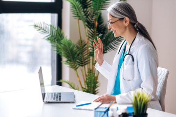 Female practitioner middle aged doctor wearing a lab coat and headset speaking by video call using...