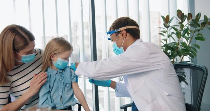 Portrait of African American young handsome happy man doctor specialist immunologist in mask injecting cute Caucasian small kid girl sitting in clinic with mom, disease prevention, vaccination concept