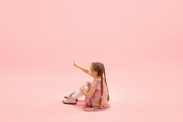 Sitting, pointing. Childhood and dream about big and famous future. Pretty longhair girl on coral pink studio background. Childhood, dreams, imagination, education, facial expression, emotions concept