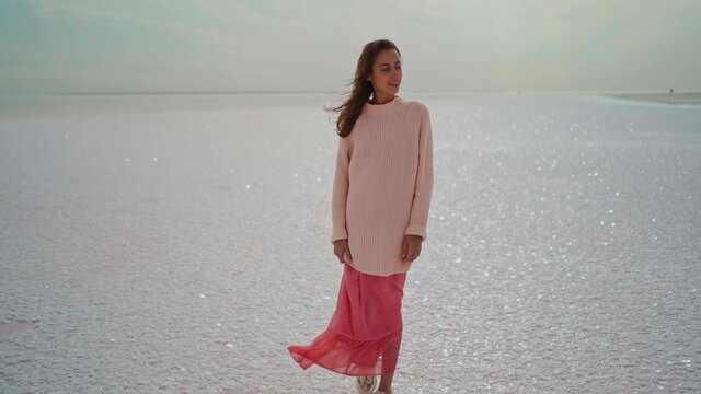 slow motion wonderful dreamy woman in big kozy sweater and pink dress standing at windy salty coastline on pink salt lake. inspired paradise picture, wellness and travel concept. nature in Ukraine