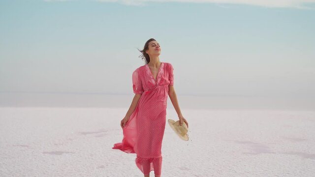 Freedom woman in blowing airness pink dress and hat standing on salty beach on pink lake. Paradise resort getaway destination at idyllic landscape