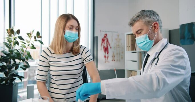 Portrait of professional handsome middle-aged doctor virologist in medical mask and white coat injecting coronavirus vaccine to Caucasian happy adult woman patient sitting in hospital, healthcare